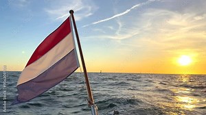 Bright colourful dutch national flag of the Netherlands flowing blowing in the wind from the stern of a sailboat during sunset sunrise at sea with blue calm sky Stock Video