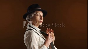 An elegant senior woman in her 60s, donning a chic fedora and holding a tie, exudes style and experience against a deep brown background. Ideal for mature fashion themes.