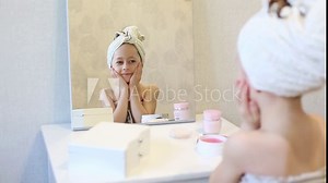 A young girl wrapped in a towel, sitting at a vanity with various skincare products, looking at her reflection in the mirror. The image captures a moment of self-care and relaxation.