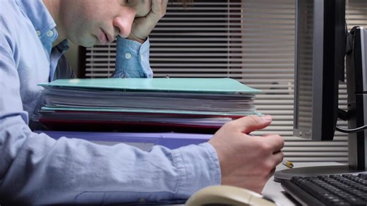 Overworked office worker feeling exhausted and overwhelmed, resting head on a large stack of documents and folders while trying to work on the computer