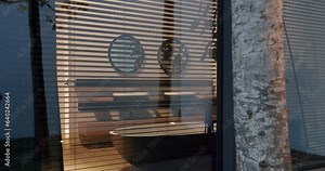 Modern cottage house, glass panoramic window with blinds, view on stylish bathroom with freestanding bath tub, washbasin, plant in pot and mirror on wall.