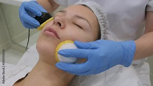 Microcurrent therapy procedure for beautiful facial skin. Young woman getting professional hardware care in modern beauty clinic. Cosmetologist's hands in gloves working with apparatus, close-up.