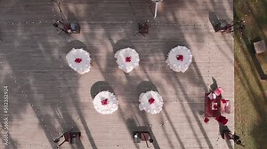 Beautiful aerial view of five round tables with white tablecloth, plates and red flowers on wooden platform with stringed garlands in middle of forest. Sunny day, trees throw oblique shadows on area.