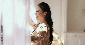 Side profile view happy Chinese woman opens curtain and admiring outside view from window at her new own or rented apartment. Welcoming new daytime in morning, start new day feel happy and optimistic