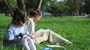 Cute happy little school children with backpacks and notebooks sitting in a grassy park outdoors. Two young schoolgirls doing homework outdoors