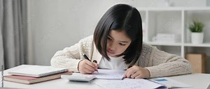 A focused Asian schoolgirl solves math homework at a tidy white desk. Concept of education and academic discipline