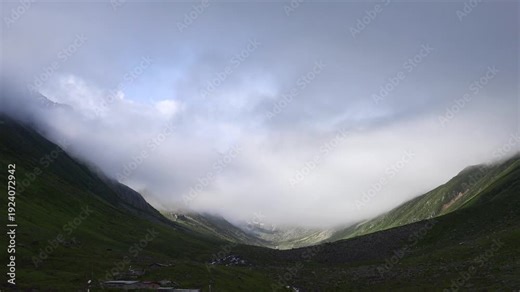 Time lapse fog and clouds disperse over high snowy mountains, revealing rugged alpine ridges. Rapid mist clearing exposes jagged peaks and glaciers above green valley slopes under shifting light.