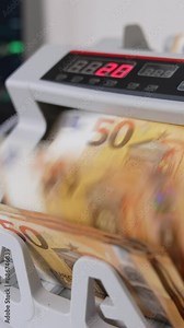 Money counting machine with a wad of fifty euro banknotes. Close up. Laptop opened at backdrop in blur. Vertical video