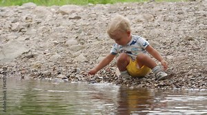 Little child playing on river bank in a summer park, 1.5 year old toddler enjoys throwing pebbles into the water. Stock Video