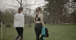 Two women enjoy a relaxing walk in the park after a workout. They're wearing fitness attire and carrying water bottles.