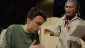 Young male actor reading script on paper and performing speech while emotional female director standing beside on stage and giving advice during theatre rehearsal