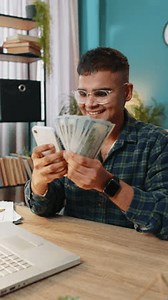 Planning family budget. Smiling Indian man counting money cash use smartphone calculate domestic bills. Hispanic guy satisfied of income and saves salary money at home office table workplace. Vertical