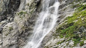 An epic view of the falling water jets of a high waterfall. The rocks on which the waterfall flows are covered with green vegetation. Slow motion. Camera movement from top to bottom