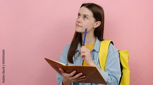 Portrait of pensive girl teen student wears shirt and yellow backpack writing down in exercise book notes, isolated on pastel pink color background wall in studio. Education in university college Stock Video