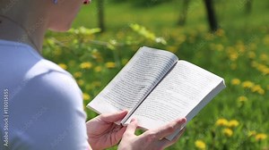 Girl reading a book sitting on a flower meadow with yellow dandelions, she turns the pages of the book
