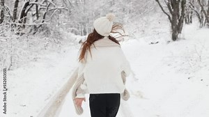 Rear view, camera follows a young long-haired woman girl running through the snowy forest of the park in a white sweater, hat and scarf, turns to the camera, smiles and extends her hand