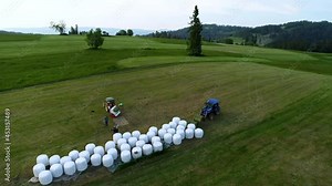 Aerial view of haymaking. Farmers working in the field. Agricultural baler machine and tractor. Fields in the mountain village. High mountain range in the background.