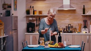 Senior woman lighting the candle waiting her husband for a romantic dinner. Elderly old wife preparing festive meal with healty food for anniversary celebration, sitting near the table in kitchen.