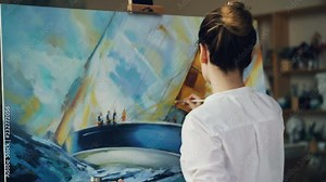Skilled artist pretty woman is painting seascape sea waves and boat with acrylic paints on canvas holding palette and brush creating masterpiece. People and arts concept.