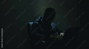 An anonymous hacker in a black hoodie cracks passwords and downloads important databases of Internet users. An Internet hacker in a black balaclava sits in a chair and steals data from a laptop