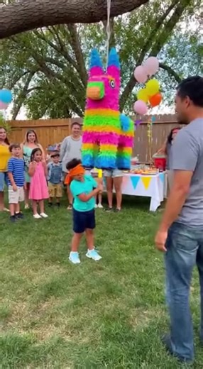 At a backyard birthday party, a blindfolded child takes a massive swing at the piñata hanging from a tree but completely misses and accidentally whacks their dad right in the stomach. The dad doubles over in pain while partygoers gasp and laugh at the unexpected hit.