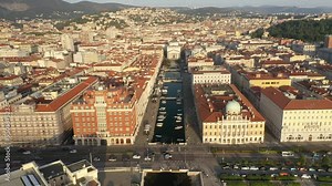 An aerial view of Trieste, Italy. The city's historic center with the Grand Canal, elegant buildings, and Piazza Unità d'Italia, surrounded by the Adriatic Sea and mountains.