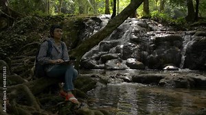Asian female ecological researcher working on digital tablet while sitting at the cascade under the shade of the trees in tropical forest. Ecosystem and environment. Conservation and scientific work.