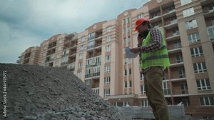 Satisfied foreman in helmet and vest at construction site talking on phone with rubble contractor and looks at waybills. Builder will check arrival building material, check shortage and call by phone.