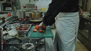 Chef cutting tomatoes and talking with female colleague when working in team during busy workday in restaurant kitchen, fish and veggies being fried in cooking pan beside