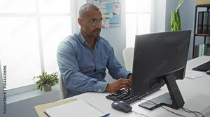 Young man working at a computer in a bright, modern office space, focused on his task with a smile suggesting productivity, surrounded by organized workspace elements and natural light.