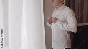 Businessman buttoning shirt near window in the morning. Stylish man groom straightens his white shirt preparing to go out. Guy get ready before date or meeting with bride fashion style weight fit