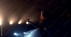 A woman with red hair is dancing on the stage. She is illuminated by the light of searchlights. She's wearing a fluffy white tutu and top. She makes smooth movements with her hands
