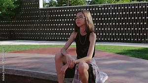 A skinny 12-year-old girl with glasses and a skateboard is sitting on a rollerdrome. Teen sports.