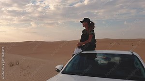 Young woman sitting on the roof of a car and looking on the epic sunset, against the background of the desert.