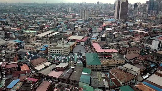 Drone aerial of Lagos Island, Nigeria, showing dense rooftops and a busy market street with a large mosque in the foreground, capturing urban life in Africa’s megacity under overcast skies.