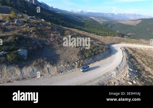 Aerial view of car driving along winding mountain road toward green forest valley. Vehicle follows curving route through rugged terrain into dense woodland landscape Stock Video Footage - Alamy