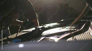 car service, a professional male mechanic in uniform repairs motor under hood with a wrench on background of sun's rays, close-up