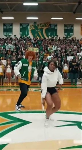 A high school pep rally is in full swing inside a brightly lit gymnasium. A group of student