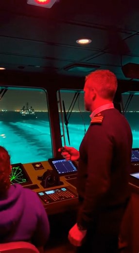 Experience an ultra-realistic internal bridge view from a cargo ship. The captain gives commands while the crew actively manages navigation systems and radar. Through the front windows, busy sea routes filled with cargo ships, tugboats, and fishing boats are visible. Unique elements include morning fog, sunlight rays, rainbow reflections, bioluminescent water, seagulls, distant lightning, and drone reflections, creating a dynamic and engaging maritime scene. Natural engine hum, radar beeps, sea 