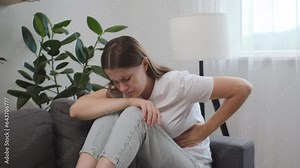Unhealthy young caucasian woman suffering from stomach ache sit on sofa. Upset brunette girl holding belly and feeling abdominal menstrual pain or bowel and digestion problems. Food poisoning concept
