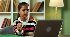 Indian Asian Cute Little Girl Sitting at Table in Modern Home, Writing in Notebook While Attending Online Class Using Laptop Computer, Focused on Homework and Learning in a Digital Education Setup