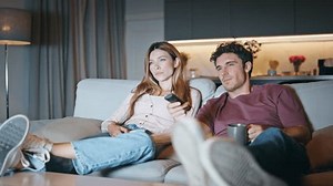 Married couple resting couch together. Smiling husband switching tv channels. Young family watching television selecting show at home interior. Two happy friends talking chilling on movie weekend