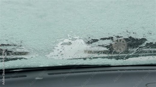 Wiper clearing ice patch on windshield leaving streaks and revealing partial view of wet road beyond