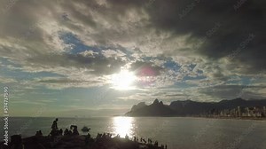 A sunset at the horizon, the sky above Ipanema Beach, reflecting off the waves of the Atlantic. Silhouettes of people, with the iconic Two Brothers Mountain in the backdrop of the Rio de Janeiro.