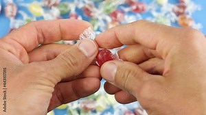 hands unfold one red candy lollipop from a transparent wrapper, on background of many colored candies, unhealthy sugar sweets, sugar addiction