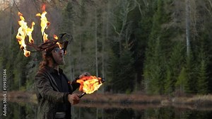 Handsome young man drinking paraffin and spitting fire using fire stick with flame during fire jamming standing near lake holding bottle in forest