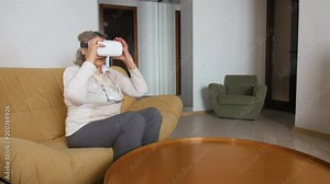 Senior Woman Sitting On The Couch Playing VR Glasses. Good-looking Senior Woman In White Using VR 360 Glasses At Home.