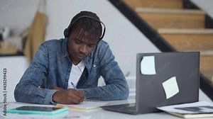 Portrait of concentrated man in earphones writing in sketchpad conferencing online in home office. Focused young African American employee handwriting ideas on remote working indoors