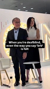 When you’re deafblind, even how you communicate “be right back” can be felt. A finger tracing from shoulder to elbow and back to the shoulder means BRB. Tactile communication keeps us connected without sight or sound. Accessibility: Rebecca stands beside a man on stage. She is wearing a white top and black skirt. Rebecca gently traces her finger down the man’s arm to communicate through touch, a form of tactile communication used by some deafblind people to share information and stay connected w