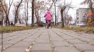 Little girl in a trendy pink outfit with matching pink bike riding her bicycle along a lane in a wooded park, learning to ride a bicycle with training wheels.